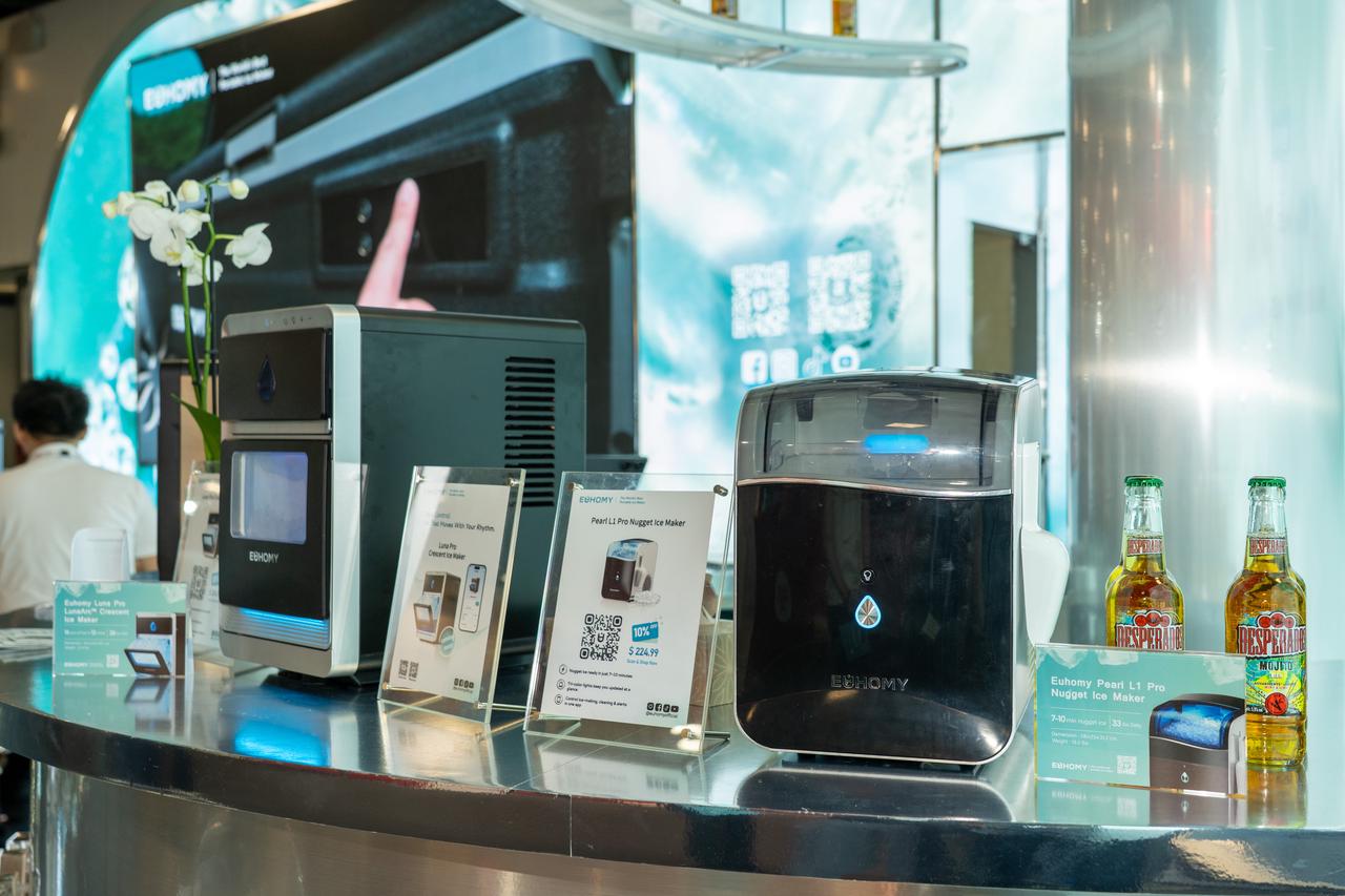 The sleek black Euhomy Pearl L1 Pro Nugget Ice Maker is displayed on a shiny counter at IFA 2025, accompanied by product specification signs and refreshing bottled beverages.