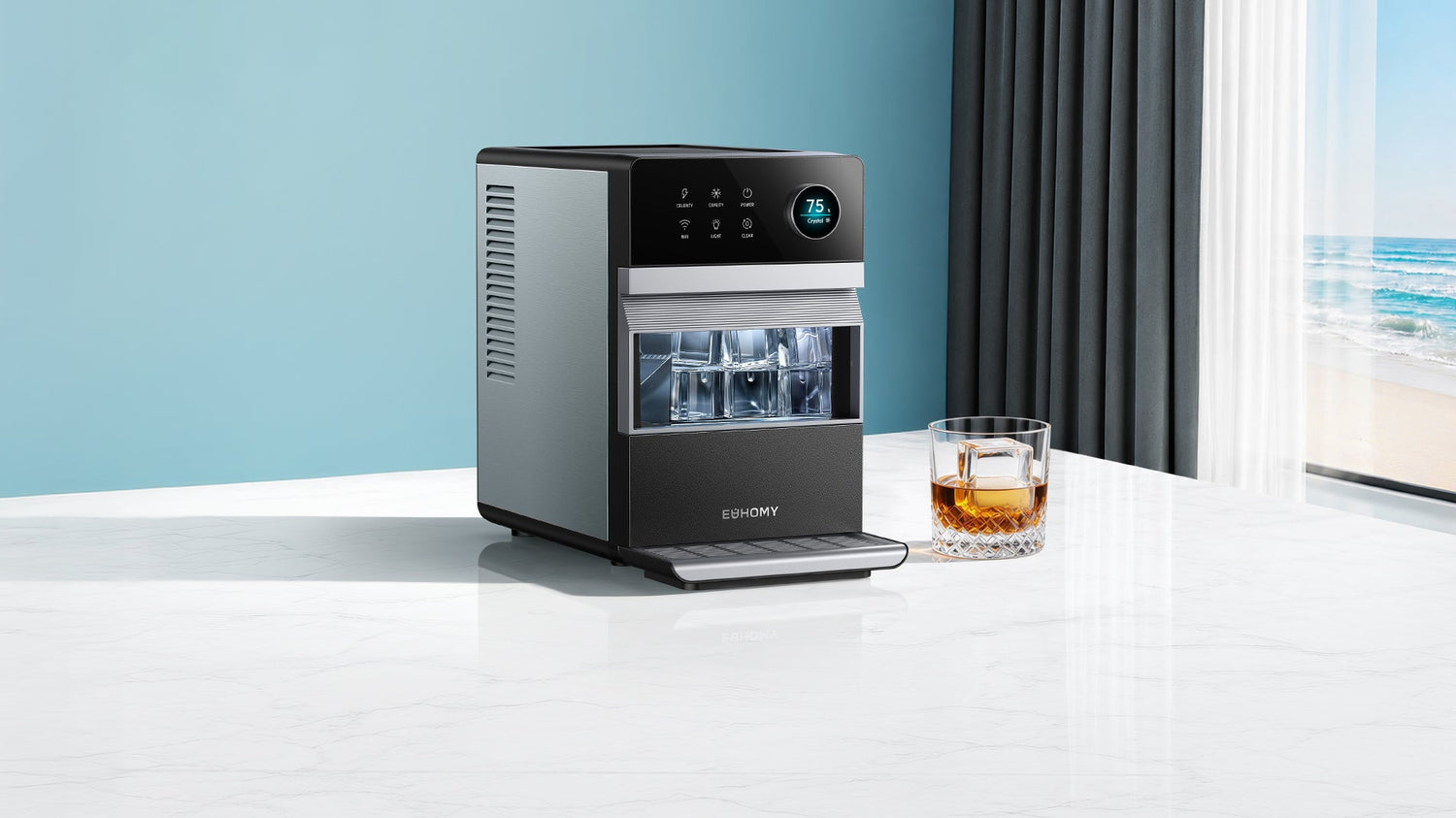 A modern, energy-efficient countertop ice maker in silver and black sits on a white marble kitchen counter next to a glass of iced whiskey, with a beach view behind.